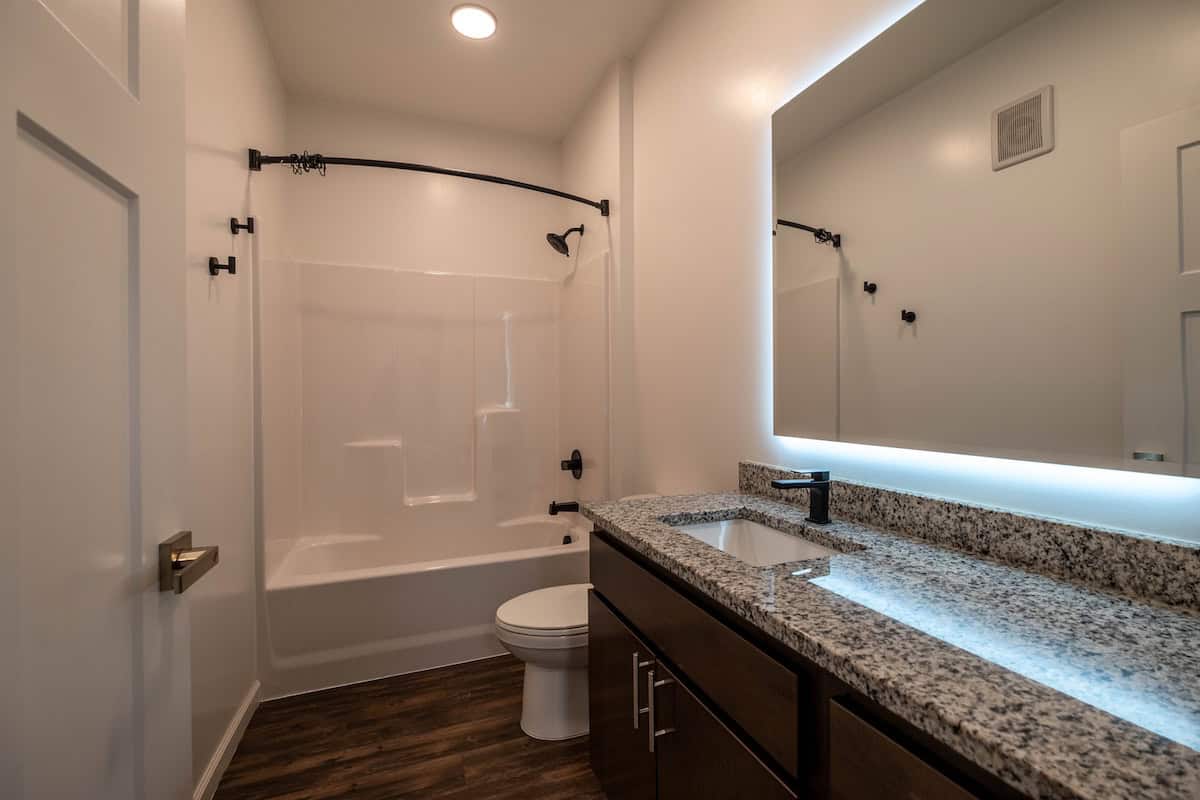 Bright bathroom featuring a shower, toilet, and granite vanity with a large illuminated mirror.